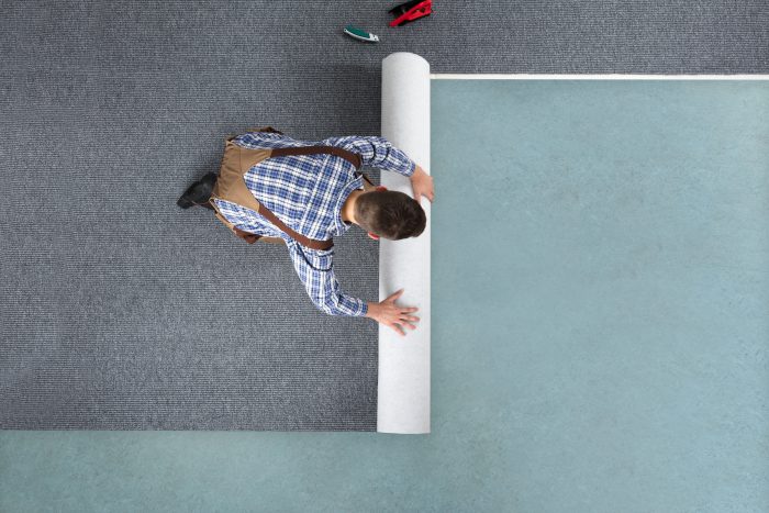 Young Male Worker In Overalls Rolling Carpet On Floor At Home